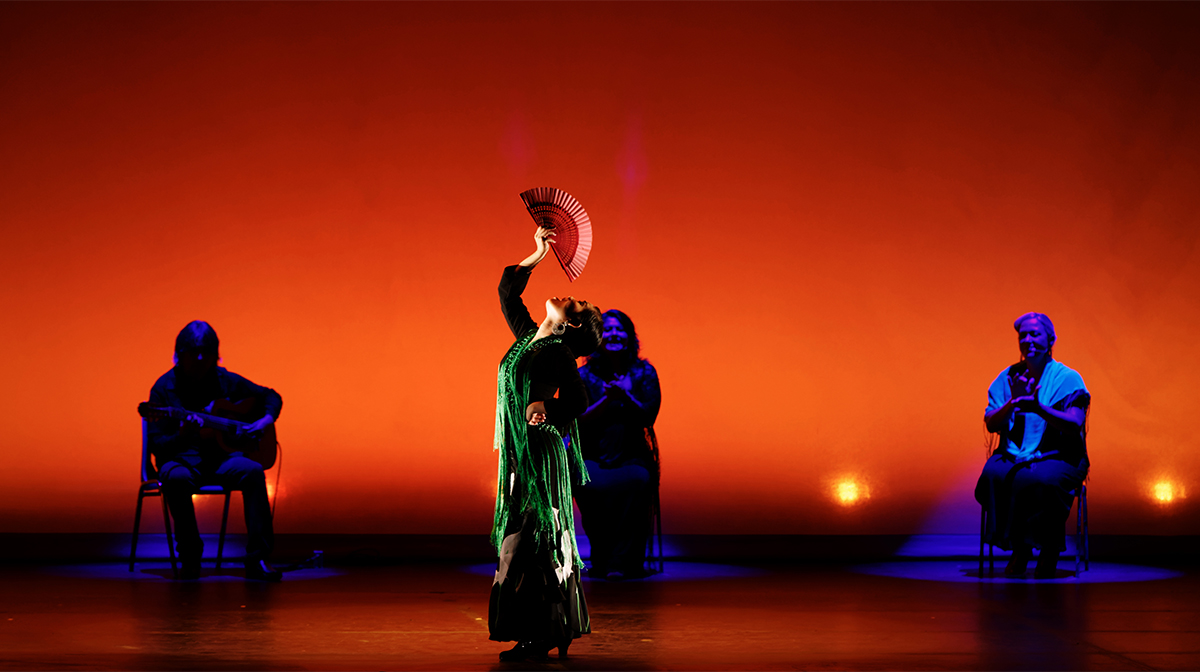 A woman in a green dress holds a flamenco fan above her. The stage is lit deep orange. Behind her three people sit clapping.